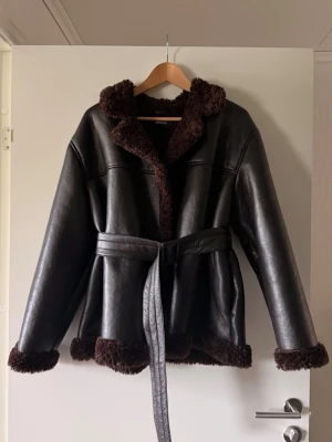 Brun Teddy Bealted jacka från Gina Tricot - Snygg brun jacka i skinnimitation med mjukt mörkbrunt teddyfoder på insidan samt vid krage, ärmslut och nederkant. Modellen har en avslappnad passform med knytskärp i midjan som ger en fin silhuett.  Jackan är endast testad.