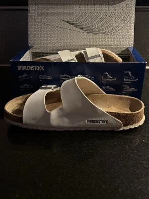 Vita Birkenstock sandaler i läder - Stilrena vita sandaler från Birkenstock med två breda remmar i läder och justerbara metallspännen. Fotbädden är i kork och mocka för extra komfort. Klassisk design som passar perfekt till sommaren.
