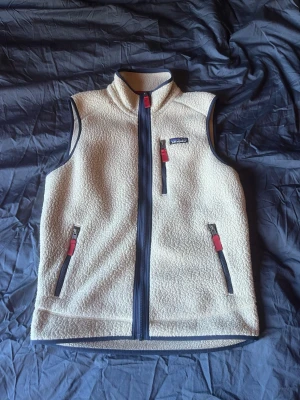 fleeceväst från Patagonia - Snygg beige fleeceväst från Patagonia med hel dragkedja framtill och tre fickor med röda detaljer. Västen har hög krage och är perfekt för lager-på-lager. Materialet är mjukt och mysigt, och västen har mörkgrå kantband. Västen är i mycket bra skick, inga fläckar eller skador osv… pris kan diskuteras!