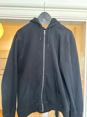 Svart zip-hoodie med fickor - En klassisk svart hoodie med dragkedja framtill och två stora fickor. Modellen har huva med dragsko och ribbade muddar vid ärmar och nederkant. Perfekt för en avslappnad och stilren look. Materialet är mjukt och bekvämt, troligtvis bomullsblandning.