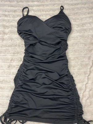 Svart minidress med dragsko - Snygg svart kortklänning med omlottdesign och smala axelband. Klänningen har snörning i sidorna som ger en rynkad effekt och gör att du kan justera längden. Perfekt för dig som gillar en figurnära passform och trendiga detaljer.