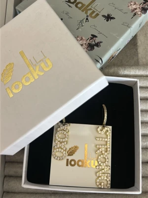 Guldiga örhängen IOAKU med text - Snygga guldiga örhängen från IOAKU med texten 'XOXO' och 'STRONG' i glittrande stenar. Örhängena har en böjd form och är täckta av små gnistrande stenar som ger en lyxig känsla. Perfekta för att göra ett statement.