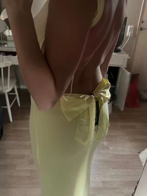 Ljusgul långklänning med rosett - Supersnygg ljusgul långklänning med öppen rygg och stor rosett bak. Klänningen har smala axelband och en tight passform som framhäver figuren. Perfekt för dig som vill sticka ut med en elegant och modern look.