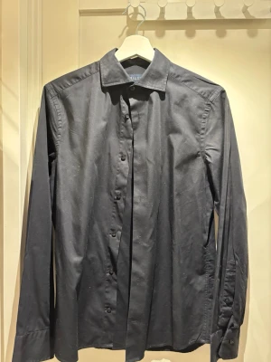 Svart skjorta från Riley | strl S - Stilren svart skjorta från Riley i slim fit-modell. Skjortan har klassisk krage, knäppning framtill och långa ärmar. Perfekt för dig som gillar en enkel och clean look. Tillverkad i mjukt bomullstyg för en bekväm känsla. Köpt på Brothers.