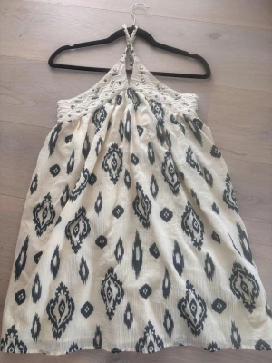 Mönstrad halterneckklänning i bomull - Snygg kortklänning med halterneck och broderade detaljer upptill. Klänningen är beige med svart mönster och har en luftig passform. Perfekt för varma dagar och sommarhäng. Tillverkad i bomull för en skön känsla.
