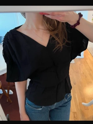 Svart v-ringad blus med korta vida ärm - Svart blus med v-ringning och korta, lösa ärmar. Modellen har markerad midja och lätt rynkade axlar för en snygg siluett. Perfekt att styla med jeans eller kjol för en trendig look. Ärmarna går att justera via snörena, beroende på hur öppna man vill ha ärmarna. Använd c.a 2 gånger så i väldigt gott skick. Storlek XS, passar strl S också.