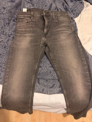 Grå jeans från Levi's - Säljer ett par grå jeans från Levi's med klassisk femficksmodell och snygga slitningar framtill. Jeansen har raka ben och normal midja. Materialet är bomullsdenim med lite stretch för extra komfort. Perfekta för en avslappnad och trendig look. Storlek 33/30