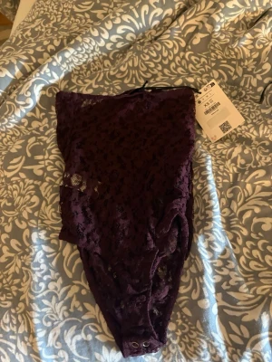 Mörklila spetsbody  - Snygg mörklila body i spets . Bodyn har hög skärning, tryckknappar i grenen och transparent spetsmaterial med blommönster. Perfekt för dig som vill ha något både bekvämt och stilrent under kläderna.