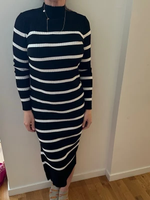 Svartvit ribbad långklänning - Snygg svart långklänning med vita horisontella ränder. Klänningen är ribbad, har lång ärm och hög hals. Materialet känns stretchigt och bekvämt, perfekt för dig som gillar en stilren och modern look.