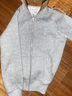 Grå zip-hoodie från Burberry - Snygg grå hoodie med dragkedja från Burberry. Klassisk huva med det ikoniska rutmönstret på insidan, två fickor framtill och diskret logga på bröstet. Perfekt för en avslappnad och trendig stil. PRIS KAN DISKUTERAS 