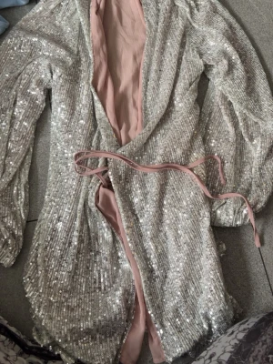 Glittrig silver omlottklänning - Snygg kort omlottklänning i silver med glittriga paljetter och ljusrosa foder. Klänningen har långa ärmar och knytband i midjan för en smickrande passform. Perfekt för dig som vill sticka ut med en festlig look.