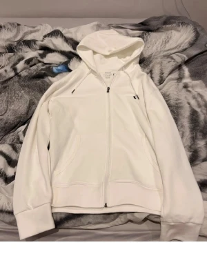Vit zip hoodie från Acne Studios - Snygg vit hoodie från Acne Studios med dragkedja framtill och två fickor. Klassisk design med huva och ribbade muddar vid ärmar och nederkant. Tillverkad i mjukt bomullsmaterial som känns skönt mot huden. Perfekt för en clean och stilren look.