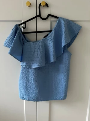 Blå offshoulder topp från Lindex - Snygg blå offshoulder topp från Lindex i strukturerat tyg. Toppen har en bred volang längs halsringningen och är ärmlös med en asymmetrisk axel. Perfekt för sommaren och skolavslutningen!
