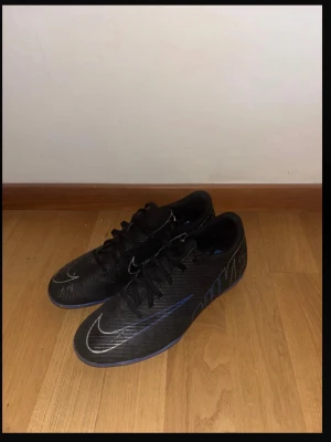 Svarta Nike  fotbollsskor - Säljer ett par svarta Nike fotbollsskor med blå detaljer och vita Nike-loggor. Skorna har snörning och är designade för att ge bra grepp och kontroll på planen. Snygg, sportig look med låg profil och syntetiskt material.