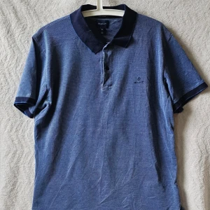Blå pikétröja från Gant - Klassisk blå pikétröja från Gant med mörkblå krage och ärmslut. Tröjan har korta ärmar, knappar vid halsen och diskret broderad logga på bröstet. Tillverkad i bomull för en skön och luftig känsla.