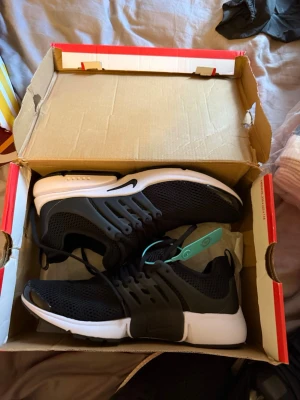 Nike Air Presto svart vita - Svarta Nike Air Presto sneakers med vit sula och svart mesh-överdel. Skorna har en sportig design med plastdetaljer vid snörningen och en flexibel, bekväm passform. Perfekta för dig som gillar stilrena och klassiska sneakers med modern känsla.