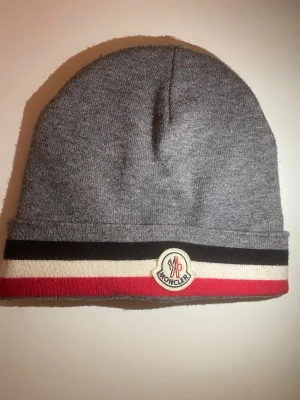 Grå Moncler mössa - Snygg grå mössa från Moncler med klassiska svarta, vita och röda ränder längst ner. Mössan har Moncler-logga framtill och är tillverkad i mjukt stickat material. Perfekt accessoar för att hålla värmen och samtidigt se trendig ut.