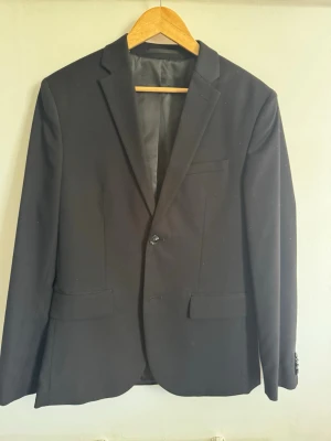 Svart klassisk kavaj - Stilren svart kavaj med smal passform och enkla slag. Kavajen har två framfickor med lock och en bröstficka. Perfekt för en dressad look och passar till skjorta eller t-shirt. Materialet är slätt och ger en snygg siluett.