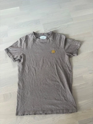 Beige t-shirt från Les Deux - Snygg beige t-shirt från Les Deux med rund hals och liten patch på bröstet. Enkel och stilren design som passar till det mesta. Tillverkad i mjuk bomull för en skön känsla hela dagen.