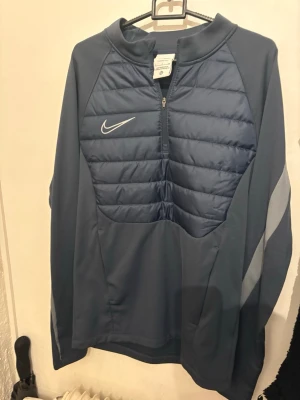 Blå Nike Therma-Fit träningströja - Snygg blå långärmad träningströja från Nike med Therma-Fit-teknologi. Tröjan har quiltad front, halv dragkedja och reflekterande detaljer på ärmarna. Perfekt för kyliga träningspass eller som överdrag. Materialet är funktionspolyester.