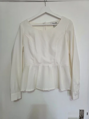 Vit peplumblus från Forever 21 - Stilren vit peplumblus från Forever 21 med långa ärmar och rund halsringning. Blusen har markerad midja och volang nertill för en feminin siluett. Tillverkad i ett lätt, vävt material och stängs med dragkedja bak.