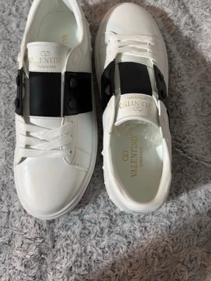 Valentino Skor - Snygga vita sneakers från Valentino Garavani med ett brett svart band över vristen. Skorna är låga och har en sula med rutmönster undertill. Tillverkade i slätt läder för en clean och exklusiv look. Köpte skorna nylisgen men de var för stora så förhoppningsvis lyckas jag sälja dem,, priset går att förhandla med