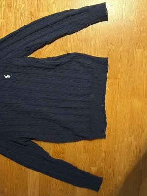 Mörkblå kabelstickad tröja Polo Ralph Lauren - Mörkblå kabelstickad tröja från Polo Ralph Lauren med klassisk vit logga broderad på bröstet. Tröjan har rund halsringning, långa ärmar och ribbade muddar vid ärmslut och nederkant. Perfekt för en stilren och avslappnad look.