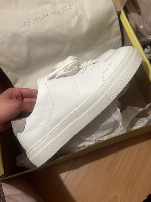 Vita sneakers från J.Lindeberg - Stilrena vita sneakers från J.Lindeberg med klassisk låg siluett. Skorna är tillverkade i slätt läder med vita snören och diskreta detaljer i grått vid hälen. Perfekta för en clean och modern look.