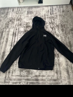 Svart hoodie från Under Armour - Snygg svart hoodie från Under Armour med dragkedja framtill och justerbar huva. Hoodien har Under Armour-logga i vitt på bröstet och är tillverkad i ett mjukt och bekvämt material. Perfekt för dig som gillar sportig och avslappnad stil. Priser kan alltid diskuteras nere.