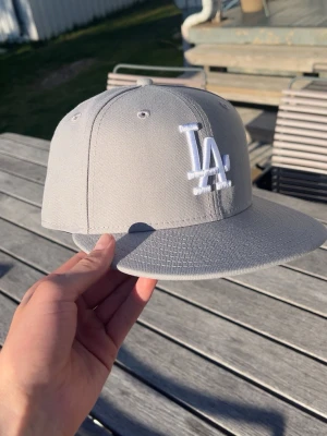 Grå LA-keps från New Era - Snygg grå snapback-keps från New Era med LA-logga broderad i vitt framtill. Klassisk rak skärm och ventilerande hål upptill. Perfekt för dig som vill ha en stilren och sportig look. Materialet är tåligt och kepsen har en strukturerad passform.                          .                                                                                                          Säljer kepsen pga att den var för stor. Jag har bara testat kepsen så det är helt ny men har dock tagit bort alla stickers. 