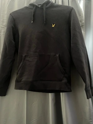 Svart hoodie från Lyle & Scott - Svart hoodie från Lyle & Scott med klassisk gul logga på bröstet. Hoodien har huva med dragsko, magficka och ribbade muddar vid ärmslut och nederkant. Perfekt för en chill och stilren look. Står xs men skulle säga strl 170