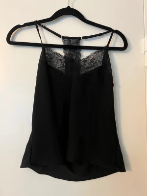 Svart linnetopp med spetsdetaljer, sov linne - Snygg svart linnetopp med tunna axelband och vacker spets upptill både fram och bak. Toppen har en lös passform och är perfekt för att skapa en stilren look. Materialet känns lätt och luftigt, och spetsen ger en extra touch av elegans. Sov linne. Bara använd ca 2 gånger 
