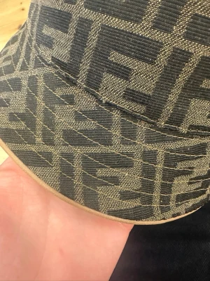 Fendi bucket hat med FF-mönster - Säljer en snygg bucket hat från Fendi med det klassiska FF-logomönstret i svart och beige. Hatten är tillverkad i ett tåligt tygmaterial och har en bred skärm som ger en cool streetwear-vibe. Perfekt accessoar för dig som gillar exklusiva designerplagg.