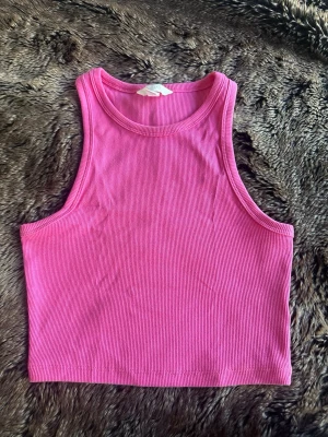 Ribbad rosa croppad linnetopp - Snygg ribbad croppad linnetopp i stark rosa färg. Toppen är ärmlös och har en klassisk rund halsringning. Perfekt för att styla med jeans eller kjol för en trendig look.