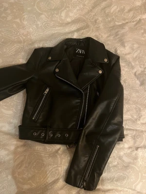 Svart skinnjacka från Zara - Snygg svart skinnjacka från Zara med klassisk bikerstil, silvriga dragkedjor och detaljer, bred krage och bälte i midjan. Jackan har två snedställda fickor med dragkedja och zip vid ärmslutet. Jackan är lite sliten på vissa ställen.
