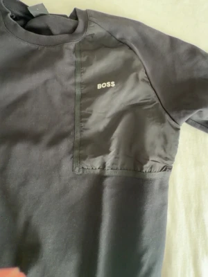 Svart t-shirt från BOSS med ficka - Snygg svart t-shirt från BOSS med en unik detalj i form av en blank bröstficka på vänster sida. T-shirten har rund halsringning och korta ärmar. Materialet är en mix av bomull och polyester vilket ger en skön känsla och modern look.
