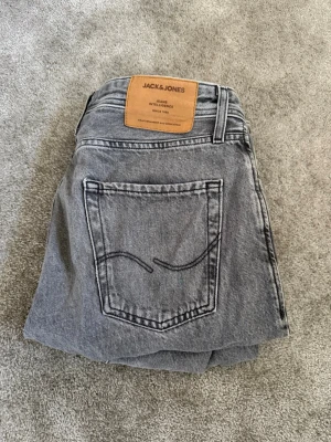 Gråa jeans från Jack & Jones - Snygga grå jeans från Jack & Jones med klassisk femficksdesign och raka ben. Jeansen har en läderpatch med logga bak i midjan och coola sömdetaljer på bakfickan. Perfekta för en avslappnad och trendig stil. 