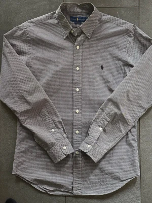 Rutig skjorta från Ralph Lauren - Snygg svartvit rutig skjorta från Ralph Lauren i slim fit-modell. Skjortan har långa ärmar, klassisk krage och knäppning framtill. Tillverkad i bomull och prydd med den ikoniska loggan på bröstet. Perfekt för en stilren look.