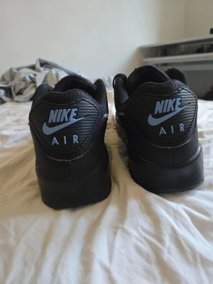 Nike Air Max 90 svart/blå sneakers - Nike Air Max 90 sneakers i svart med ljusblå detaljer och logga. Klassisk chunky sula med synlig Air-enhet, ovandel i mesh och syntetmaterial. Swoosh och AIR MAX-text i blått på sidan. Perfekta för dig som gillar streetwear och sportig stil. Orginal skor från Nike som är testad någångång, så som helt nya