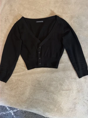 Svart cropped kofta från Brandy Melville - Svart, ribbstickad cropped kofta från Brandy Melville med v-ringning, trekvartsarmar och knäppning framtill. Långa ärmar och bred ribbad mudd vid midjan ger en snygg passform. Perfekt att styla över linne eller t-shirt.