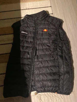 Svart dunväst från Ellesse - Stilren svart dunväst från Ellesse med broderad logga på bröstet. Västen har hög krage och quiltad design som ger en sportig look. Perfekt att slänga över en hoodie eller tröja för extra värme.
