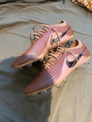 Nike rosa fotbollsskor med metallicswoosh - Säljer ett par rosa Nike fotbollsskor med metallicfärgad swoosh på sidan. Skorna har snörning och en modern design med vågformade detaljer på ovandelen. Yttersulan har fasta dobbar för bra grepp på planen.