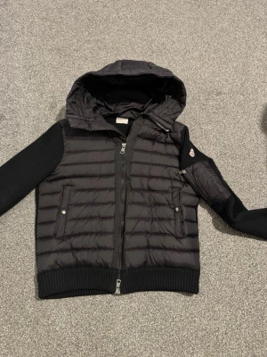 Moncler cardigan  - Snygg svart pufferjacka från Moncler med huva och stickade ärmar. Jackan har två fickor framtill med knappar och en extra ficka med dragkedja på ena ärmen. Klassisk Moncler-logga på ärmen och ribbade muddar.