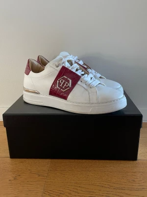 Philipp Plein vita och vinröda sneakers - Snygga vita sneakers från Philipp Plein med vinröda detaljer i croco-mönstrat läder och silverfärgad logga på sidan. Skorna har lågt skaft, vita snören och en chunky sula. Perfekta för dig som vill sticka ut med exklusiv streetstyle.