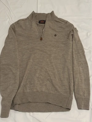 Beige halvzip-tröja Morris - Snygg beige långärmad tröja från Morris med halvzip och liten broderad logga på bröstet. Tröjan har ribbade muddar vid ärmslut och nederkant, perfekt för lager på lager. Materialet ser ut att vara mjukt och tunt, troligen bomull eller ullmix.