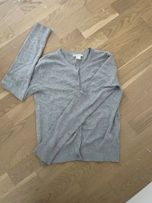 Grå basic kofta med knappar - En enkel och stilren grå kofta från H&M Basic med rund halsringning och knäppning framtill. Koftan har långa ärmar och ribbade muddar vid ärmslut och nederkant. Perfekt att slänga över en t-shirt eller linne.
