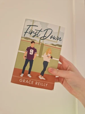 First Down av Grace Reilly - En färgglad pocketbok med titeln 'First Down' av Grace Reilly. Omslaget visar en tecknad kille i lila tröja och en tjej med blont hår på en fotbollsplan. Boken har en modern, ungdomlig design och handlar om kärlek och amerikansk fotboll. I begagnat skick