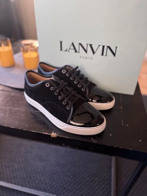 Lanvin DBB1 Cap Toe Sneaker – Svart | Strl 43 | Fabriksnya Med Box - Lanvin DBB1 Cap Toe i klassisk svart — ikonisk fransk lyx med patent läder finish och signatur cap toe design. ⚠️ Storleksguide: Denna modell är stor i storleken. Beställ en storlek mindre än vanligt. Bär du vanligtvis 44 väljer du 43. Fabriksnya, aldrig använda. Medföljer: ✅ Originallåda ✅ Dustbag ✅ Kvitto Storlek 43. Kan även köpas via vår hemsida www.nordens.store med Klarna & Paypal 😊 Vi tar även emot byten och uppgraderingar! 🔄 📦 Spårad frakt, väl förpackad.