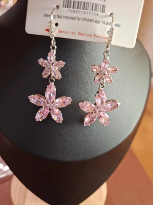 Rosa blommiga hängande örhängen - Säljer ett par glittriga örhängen i form av dubbla blommor med ljusrosa stenar. Örhängena har silverfärgade detaljer och gnistrar snyggt i ljuset. Perfekta för dig som vill ha något extra till din look.
