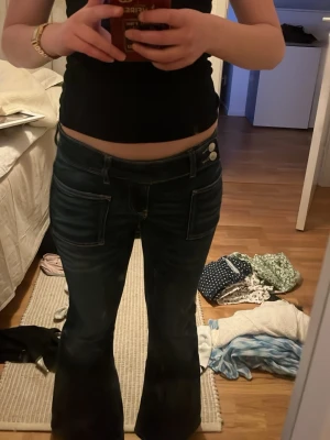 Mörkblå bootcut jeans Gina Tricot - Säljer ett par mörkblå utsvängda jeans från Gina Tricot med två stora fickor fram och dubbla knappar i midjan. Jeansen har en snygg retrostil och är tillverkade i klassisk denim. Perfekta för dig som gillar trendiga och bekväma byxor!